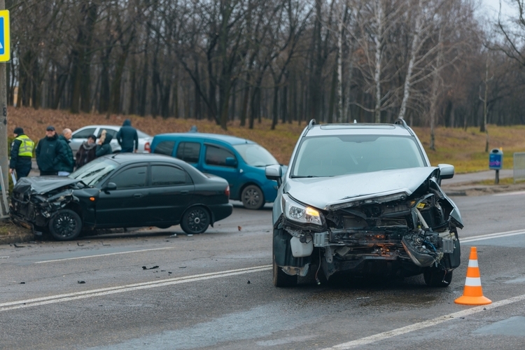 A silver car with significant front-end damage and a black car with a crushed hood are stopped in the middle of a road following a collision. A group of people stands nearby as an orange traffic cone marks the scene.