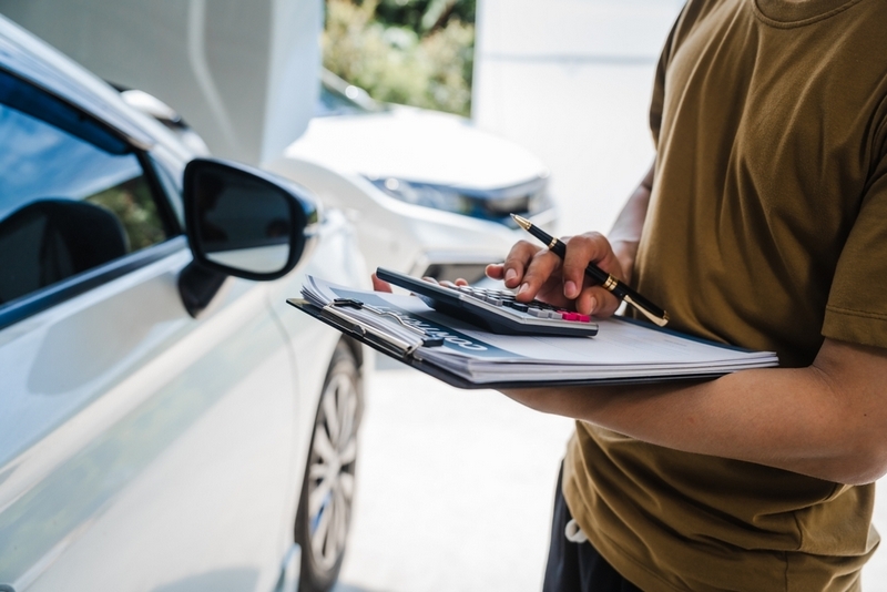 A person holding a clipboard and pen stands next to a white car, filling out paperwork outdoors.